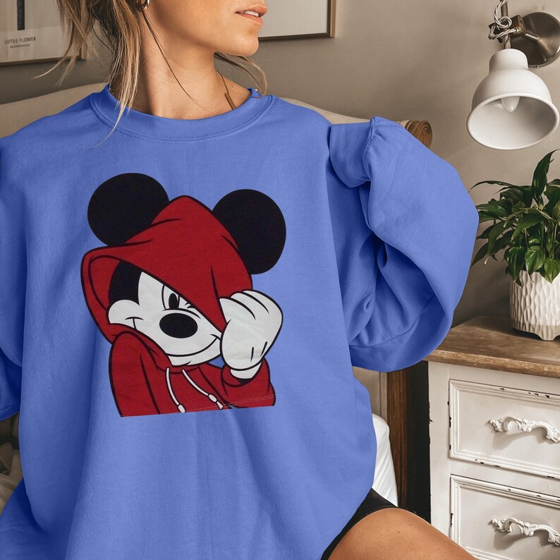 May include: A periwinkle blue sweatshirt featuring a graphic of Mickey Mouse wearing a red hoodie. Mickey's black ears are visible above the hood. The graphic is centered on the front of the long-sleeved top.