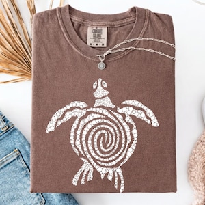 May include: A brown Comfort Colors t-shirt with a white turtle design. The turtle has a spiral shell and is made of small white circles. A silver necklace with a pendant is draped across the shirt.