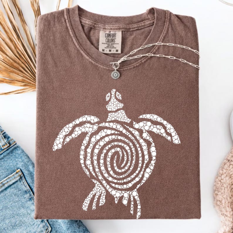 May include: A brown Comfort Colors t-shirt with a white turtle design. The turtle has a spiral shell and is made of small white circles. A silver necklace with a pendant is draped across the shirt.