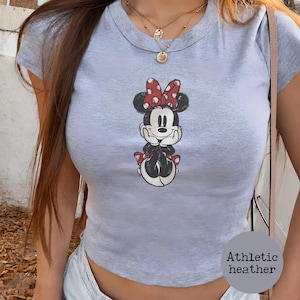 May include: A light gray athletic heather crop top featuring a Minnie Mouse graphic. The cartoon character is depicted in black, white, and red, with a red bow and polka dots. The top has a round neckline and short sleeves.