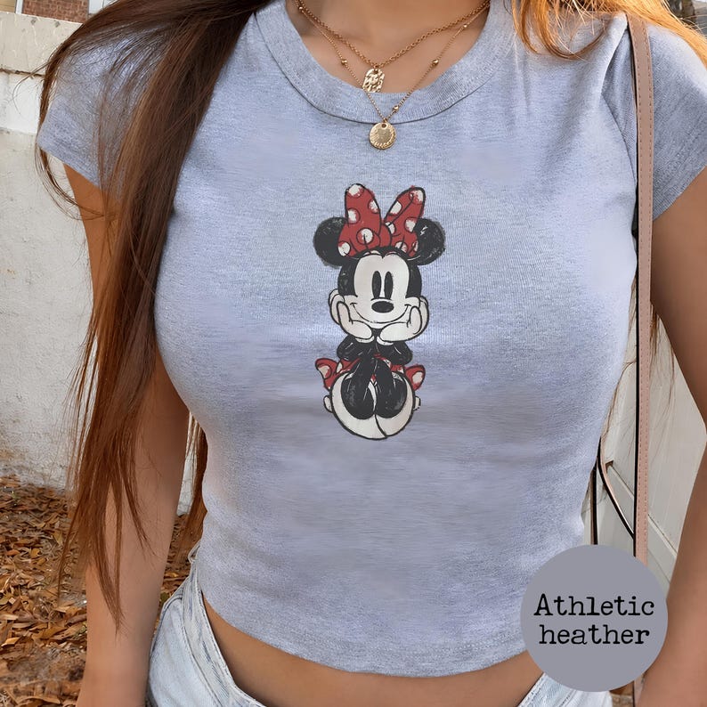 May include: A light gray athletic heather crop top featuring a Minnie Mouse graphic. The cartoon character is depicted in black, white, and red, with a red bow and polka dots. The top has a round neckline and short sleeves.