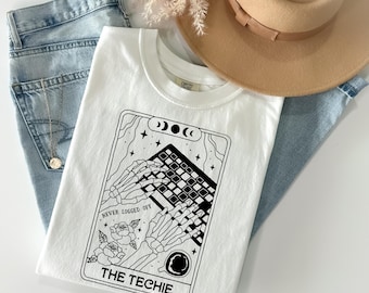 Könnte beinhalten: Weißes T-Shirt mit einer schwarzen Grafik von Skeletthänden, die auf einer Tastatur tippen, mit dem Text "NEVER LOGGED OFF" und "THE TECHIE". Das Shirt liegt auf einer blauen Jeans, mit einem beigen Hut und einer goldenen Halskette.