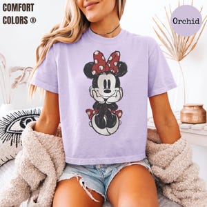 May include: A light purple t-shirt featuring a distressed graphic of Minnie Mouse with a red polka dot bow. The shirt has short sleeves and the text "COMFORT COLORS" is visible in the upper left corner. The color of the shirt is "Orchid".