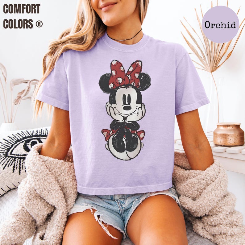 May include: A light purple t-shirt featuring a distressed graphic of Minnie Mouse with a red polka dot bow. The shirt has short sleeves and the text "COMFORT COLORS" is visible in the upper left corner. The color of the shirt is "Orchid".