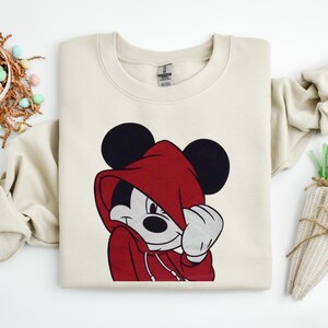 May include: Cream-colored sweatshirt featuring a graphic of Mickey Mouse in a red hooded sweatshirt. Mickey's black ears are visible above the hood. The design is centered on the front of the garment, with a white drawstring and a white hand.