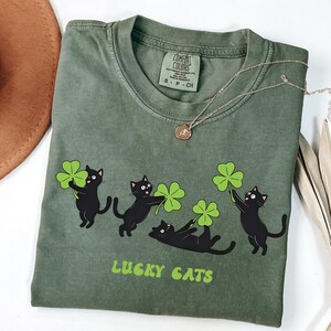 May include: Olive green t-shirt featuring a graphic of four black cats holding green shamrocks. The text "LUCKY CATS" is printed in green below the cats. A gold necklace with a pendant is draped across the shirt.