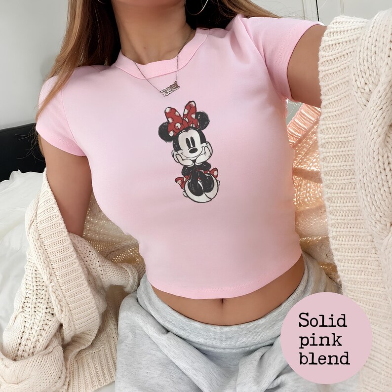 May include: A light pink crop top featuring a graphic of Minnie Mouse with a red bow. The top has short sleeves and a crew neck. The image also includes a light pink circle with the words "Solid pink blend".