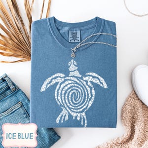 May include: Ice blue Comfort Colors t-shirt with a white turtle graphic. The shirt has a crew neck and short sleeves. A silver necklace with a turtle pendant is draped over the shirt. The shirt is folded on a white surface.