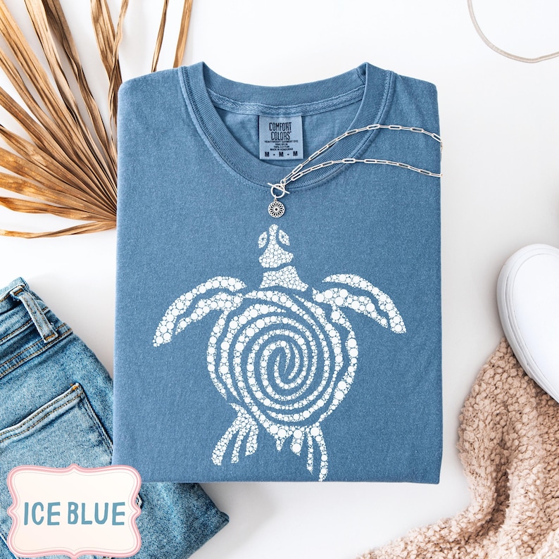 May include: Ice blue Comfort Colors t-shirt with a white turtle graphic. The shirt has a crew neck and short sleeves. A silver necklace with a turtle pendant is draped over the shirt. The shirt is folded on a white surface.