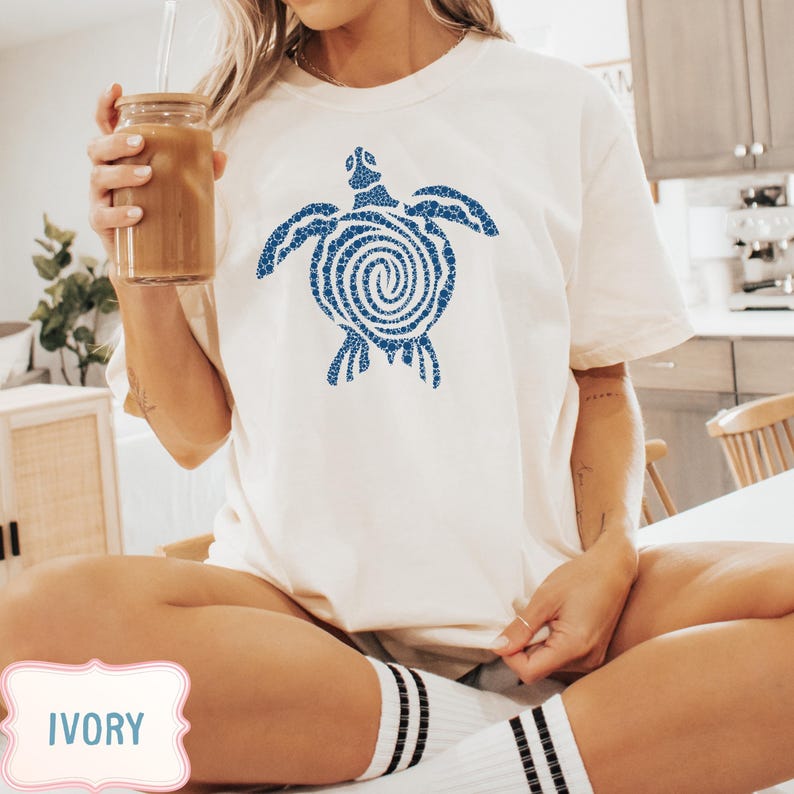 May include: Ivory-colored t-shirt featuring a blue turtle design. The turtle graphic is centered on the shirt and has a spiral pattern in its shell. The shirt is paired with white socks with black stripes.