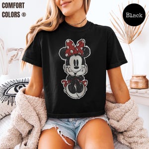 May include: Black t-shirt featuring a distressed graphic of Minnie Mouse with a red and white polka dot bow. The shirt has the words "COMFORT COLORS" in white text on the upper left. A black circle with the word "Black" is on the upper right.