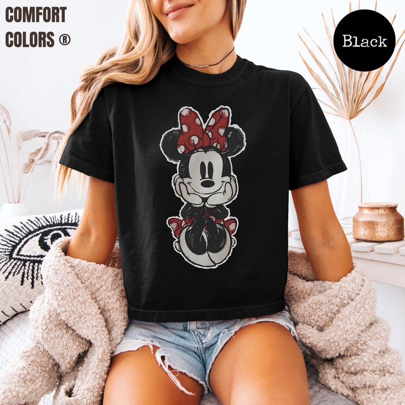 May include: Black t-shirt featuring a distressed graphic of Minnie Mouse with a red and white polka dot bow. The shirt has the words "COMFORT COLORS" in white text on the upper left. A black circle with the word "Black" is on the upper right.