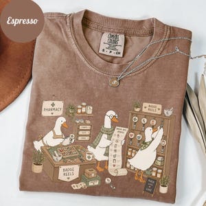 May include: Espresso-colored Comfort Colors t-shirt featuring a pharmacy scene with cartoon geese. The geese are depicted working behind a counter, with the text "Pharmacy" and "Badge Reels" visible. A silver necklace is draped over the shirt.