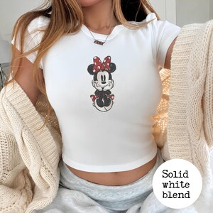 May include: White short-sleeved t-shirt featuring a Minnie Mouse graphic. The cartoon character is depicted in black, red, and white. The shirt has a round neckline and a fitted silhouette. The text "Solid white blend" is in a white circle.