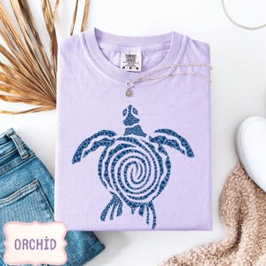May include: A lilac Comfort Colors t-shirt with a navy blue turtle design. The turtle graphic is made of small circles. A silver necklace with a small pendant rests on the shirt. The word "ORCHID" is in a pink frame.