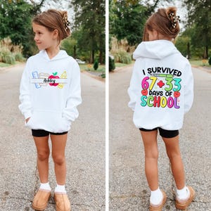 May include: A white hoodie featuring the name "Ashley" and school-related illustrations on the front. The back displays the text "I SURVIVED 67+33 DAYS OF SCHOOL" in vibrant colours. The hoodie is worn by a young person.