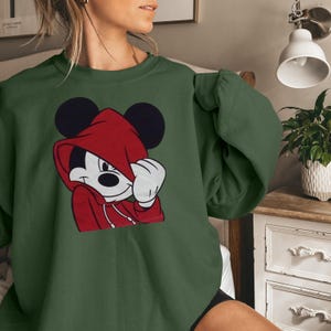 May include: Green sweatshirt featuring a graphic of Mickey Mouse in a red hoodie. The cartoon character is depicted with a white face, black ears, and a red hoodie. The sweatshirt is a solid green color.