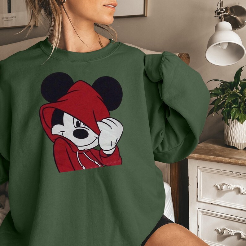 May include: Green sweatshirt featuring a graphic of Mickey Mouse in a red hoodie. The cartoon character is depicted with a white face, black ears, and a red hoodie. The sweatshirt is a solid green color.