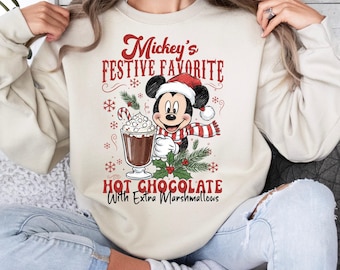 May include: Beige sweatshirt featuring a festive graphic of Mickey Mouse in a Santa hat and scarf, holding a cup of hot chocolate with marshmallows. The text reads "Mickey's Festive Favorite" and "Hot Chocolate With Extra Marshmallows".
