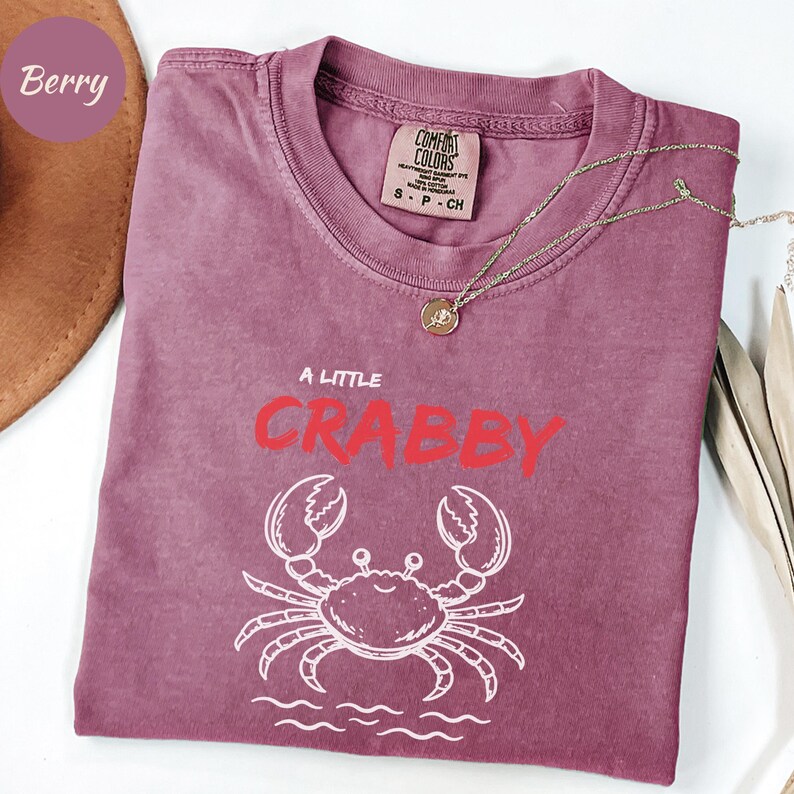 May include: Berry-colored t-shirt with the phrase "A Little Crabby" in red and a white crab graphic. The shirt has a crew neck and short sleeves. A gold necklace with a pendant is draped across the shirt.