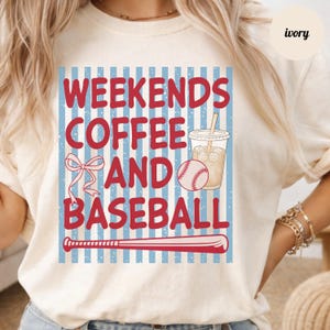 May include: Ivory t-shirt with a red graphic design that reads "Weekends Coffee and Baseball". The design includes a baseball, a coffee drink, a bow, and a bat, set against a blue and white striped background.