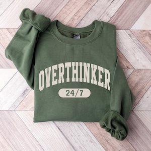Pode incluir: Camisola verde escuro com a palavra "OVERTHINKER" em letras grandes, brancas. Abaixo, o texto "24/7" num retângulo arredondado branco. A camisola está dobrada sobre uma superfície de madeira castanha clara.