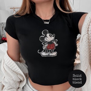K&ouml;nnte beinhalten: Schwarzes Crop-Top mit einem verblichenen Mickey Mouse-Motiv in Schwarz, Wei&szlig; und Rot. Die Zeichentrickfigur ist in einer klassischen Pose dargestellt. Das Top hat kurze &Auml;rmel und einen Rundhalsausschnitt. Der Text "Solid black blend" befindet sich unten rechts.