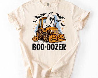 Peut inclure: T-shirt crème avec un motif d'Halloween. Le dessin représente un fantôme conduisant un bulldozer orange avec des lames en forme de citrouille, des chauves-souris et le texte "BOO-DOZER" en noir.