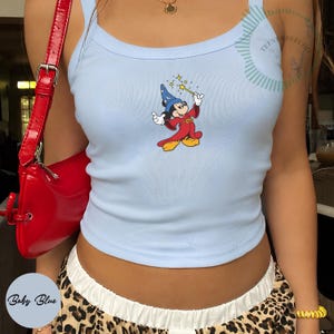 May include: A light blue ribbed tank top featuring a graphic of a cartoon mouse wearing a blue wizard hat and a red robe, holding a yellow magic wand with sparkling stars. The top has a scoop neckline and is paired with a red shoulder bag.