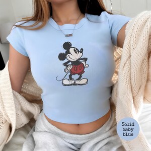 K&ouml;nnte beinhalten: Hellblaues Crop-Top mit einem klassischen Mickey Mouse-Motiv. Das Shirt hat kurze &Auml;rmel und einen runden Ausschnitt. Die Grafik zeigt Mickey Mouse in seinen roten Shorts und wei&szlig;en Handschuhen. Das Shirt ist einfarbig hellblau.