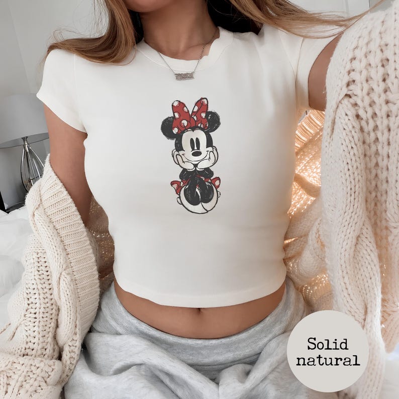 May include: Cream-colored crop top featuring a Minnie Mouse graphic. The cartoon character is depicted in black, red, and white. The top has short sleeves and a round neckline. The words "Solid natural" are in a circle in the bottom right corner.