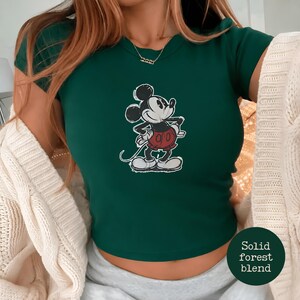 K&ouml;nnte beinhalten: Ein dunkelgr&uuml;nes T-Shirt mit einem klassischen Mickey Mouse-Motiv. Die Zeichentrickfigur ist in Schwarz, Wei&szlig; und Rot dargestellt. Das Shirt hat einen Rundhalsausschnitt und kurze &Auml;rmel. Der Text "Solid forest blend" befindet sich in einem Kreis unten rechts.