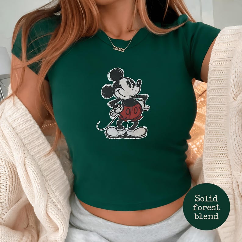 K&ouml;nnte beinhalten: Ein dunkelgr&uuml;nes T-Shirt mit einem klassischen Mickey Mouse-Motiv. Die Zeichentrickfigur ist in Schwarz, Wei&szlig; und Rot dargestellt. Das Shirt hat einen Rundhalsausschnitt und kurze &Auml;rmel. Der Text "Solid forest blend" befindet sich in einem Kreis unten rechts.