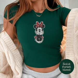 May include: A forest green t-shirt featuring a Minnie Mouse graphic with a red bow and polka dots. The shirt has short sleeves and a crew neck. The text "Solid forest blend" is in a circle on the bottom right.
