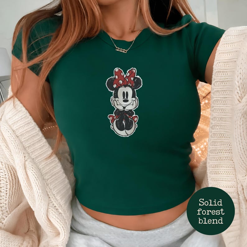 May include: A forest green t-shirt featuring a Minnie Mouse graphic with a red bow and polka dots. The shirt has short sleeves and a crew neck. The text "Solid forest blend" is in a circle on the bottom right.