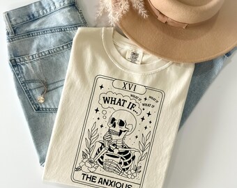 May include: Cream-coloured t-shirt with a tarot card design featuring a skeleton, the text "WHAT IF...", and "THE ANXIOUS". The shirt is styled with light blue jeans, a tan hat, and a gold necklace.