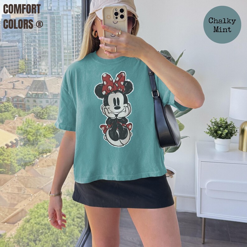 May include: A teal Comfort Colors t-shirt featuring a Minnie Mouse graphic with a red bow. The shirt has short sleeves and a relaxed fit. The words "Comfort Colors" are printed in white on the upper left. A round label says "Chalky Mint".