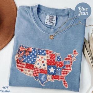 May include: A blue jean coloured t-shirt featuring a patchwork design of the United States map. The map is made of red, white, and blue patterned fabric. A gold necklace with a pendant is draped on the shirt. The text "Blue Jean" is in a blue circle.
