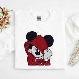 May include: White sweatshirt featuring a graphic of Mickey Mouse wearing a red hoodie. The cartoon character is smiling and covering part of his face with his hand. The sweatshirt has a crew neck and long sleeves.