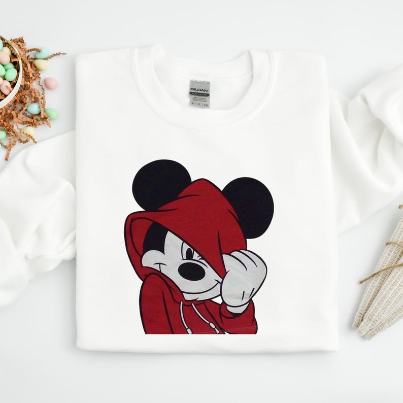 May include: White sweatshirt featuring a graphic of Mickey Mouse wearing a red hoodie. The cartoon character is smiling and covering part of his face with his hand. The sweatshirt has a crew neck and long sleeves.