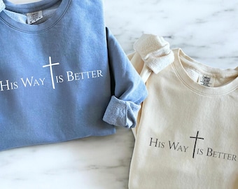 May include: Two crewneck sweatshirts, one light blue and one cream. Both feature the text "His Way Is Better" with a cross graphic. The light blue sweatshirt has rolled-up sleeves. The sweatshirts are made of a soft material.