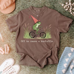 Könnte beinhalten: Braunes T-Shirt mit einer Illustration eines grünen Frosches auf einem Fahrrad, der einen rot-weißen Hut trägt. Der Text "Off to cause a kerfuffle" ist darunter gedruckt.