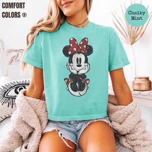 May include: A turquoise t-shirt featuring a cartoon illustration of Minnie Mouse with a red polka dot bow. The shirt has the words "Comfort Colors" printed on the top left. The words "Chalky Mint" are printed on the top right.
