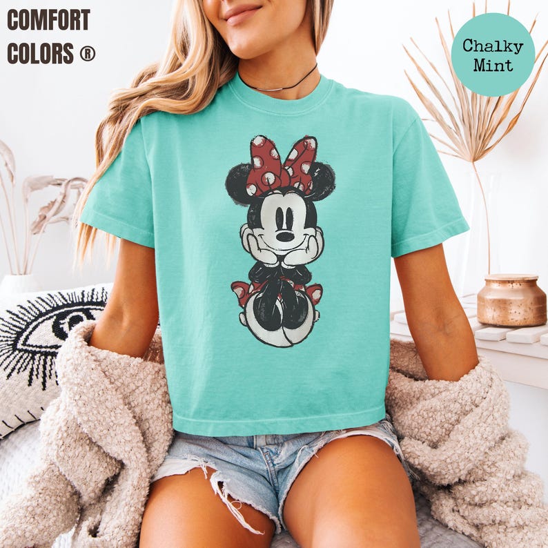 May include: A turquoise t-shirt featuring a cartoon illustration of Minnie Mouse with a red polka dot bow. The shirt has the words "Comfort Colors" printed on the top left. The words "Chalky Mint" are printed on the top right.