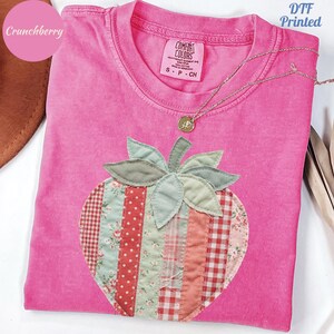 May include: A pink Comfort Colors t-shirt featuring a patchwork strawberry design. The strawberry is made of various patterned fabrics in red, pink, and green. A gold necklace with a pendant rests on the shirt. The text "DTF Printed" is visible.