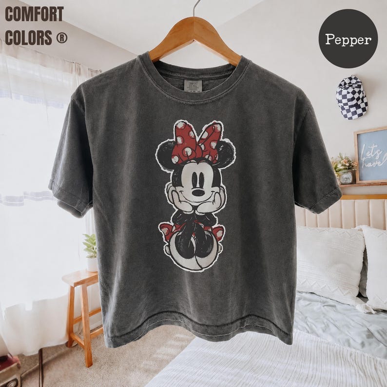 May include: A dark gray Comfort Colors t-shirt featuring a Minnie Mouse graphic. The graphic shows Minnie Mouse with a red polka dot bow and red accents. The shirt is hanging on a wooden hanger.