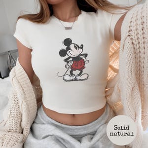 K&ouml;nnte beinhalten: Cremefarbenes Crop-Top mit einem Mickey Mouse-Motiv. Die Cartoonfigur ist in Schwarz, Rot und Wei&szlig; dargestellt. Das Oberteil hat kurze &Auml;rmel und einen runden Ausschnitt. Der Text "Solid natural" befindet sich in einem Kreis unten.