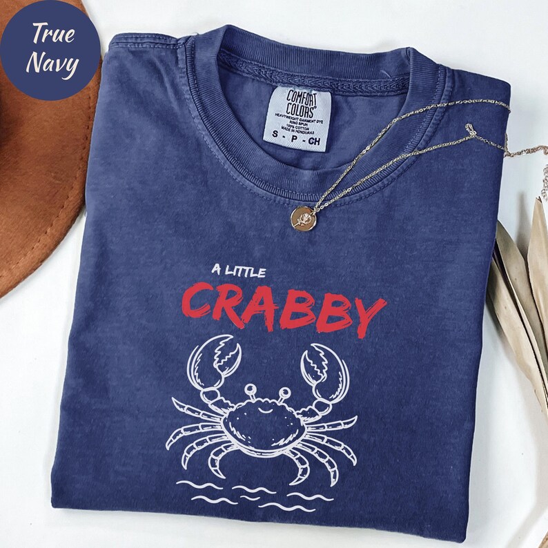 May include: A navy blue t-shirt with the text "A Little Crabby" in red and a white crab graphic. The shirt has a crew neck and short sleeves. A gold necklace with a pendant is draped across the shirt.