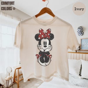 May include: Ivory-colored t-shirt featuring a distressed graphic of Minnie Mouse with a red polka-dot bow. The shirt is hanging on a wooden hanger. The text "COMFORT COLORS" is visible in the upper left corner.