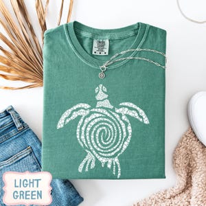 May include: A light green t-shirt with a white turtle design. The shirt is folded and has a silver necklace with a pendant. The words "LIGHT GREEN" are in a pink and white label in the bottom left corner. A pair of blue jeans and a white shoe are also visible.